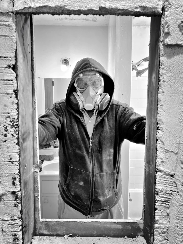 A man with a respirator, goggled, and hoodie stands behind an opening in a bathroom wall.